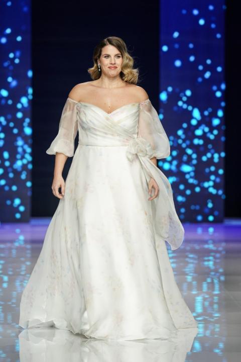 Abito da sposa curvy DELPHINE, scollo a cuore e maniche trasparenti, su passerella con sfondo blu.