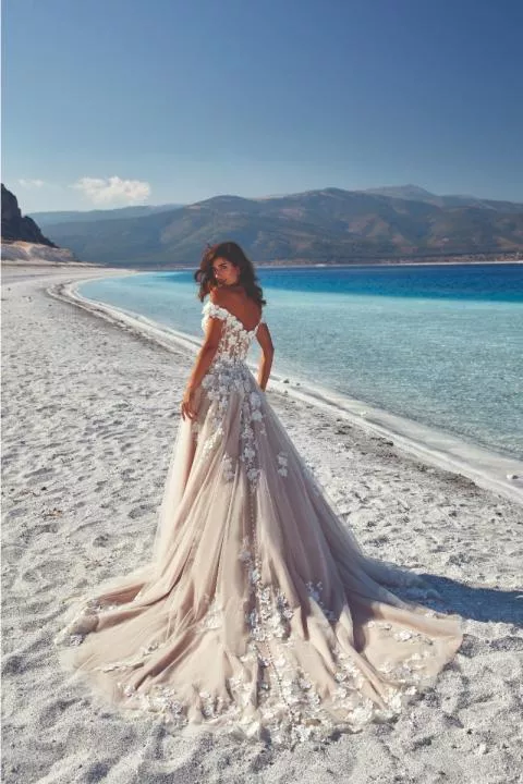 Abito da sposa Lima Eddy K in spiaggia, dettagli floreali, mare e monti sullo sfondo.