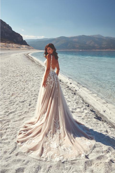 Abito da sposa FRANCA di Eddy K su spiaggia, schiena scoperta e dettagli floreali, mare e montagne sullo sfondo.