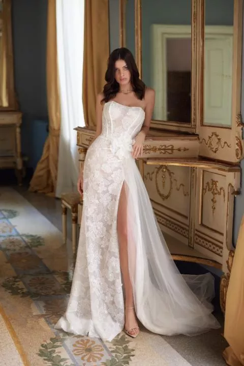Abito da sposa PEARL - MARK LOREN in pizzo bianco con spacco alto, ambientato in una stanza elegante.