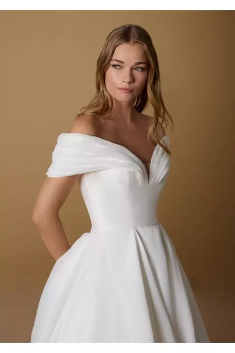 Abito da sposa PRISCA bianco con scollo a cuore e spalle scoperte, sfondo beige.