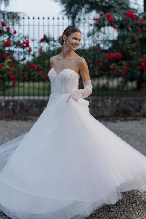 Abito da sposa JACLYN di SPOSAE, design romantico con corpetto in pizzo, sfondo floreale.