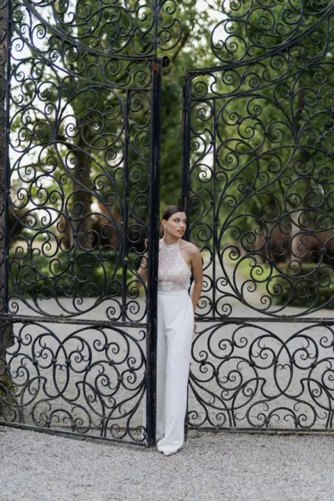 Donna in abito da sposa ADRIENNE di Sposae Atelier davanti a un cancello ornato, ambiente elegante e naturale.