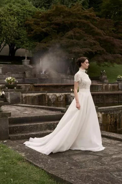 Abito da sposa Beatrice in pizzo e chiffon, collezione Romantica, in giardino con fontana sullo sfondo.
