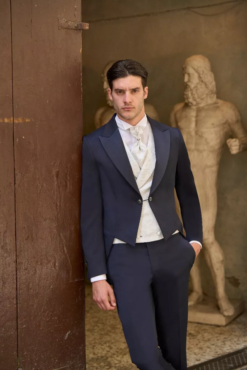 Abito da cerimonia Rocchini 2, uomo in giacca blu e gilet bianco, sfondo con statue antiche.