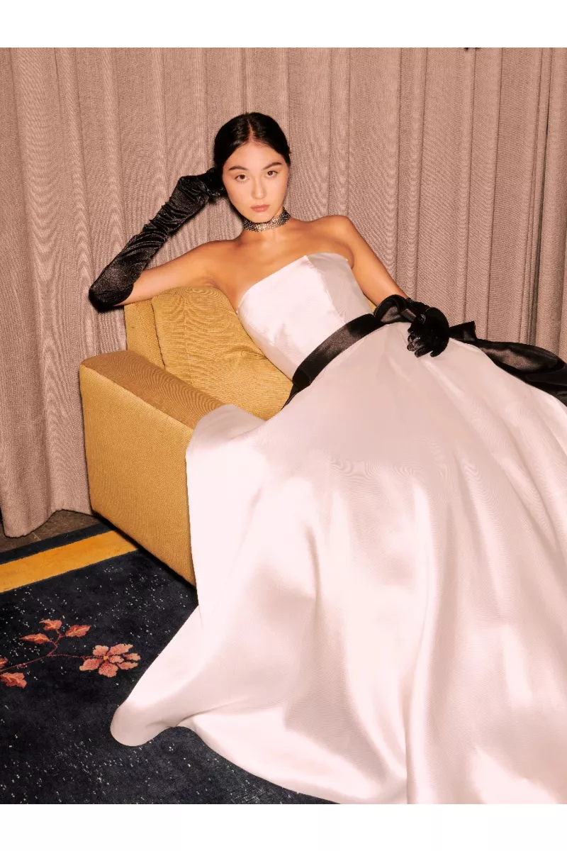 Abito da sposa Marilù Sposae Essential, elegante con guanti neri e cintura, su divano giallo.