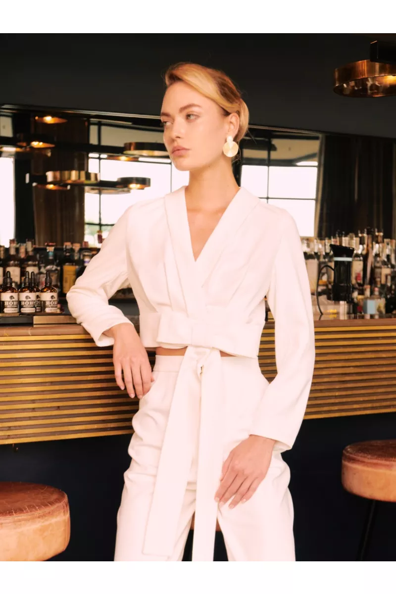 Completo bianco elegante Livorno Sposae Essential, donna in un bar moderno, orecchini dorati, atmosfera sofisticata.