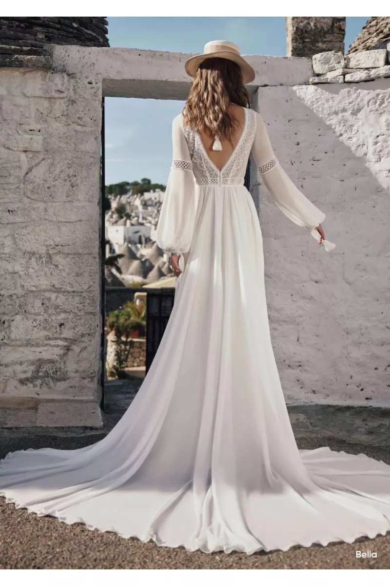 Abito da sposa boho Bella Ariamo, maniche lunghe in pizzo, sfondo architettura bianca.