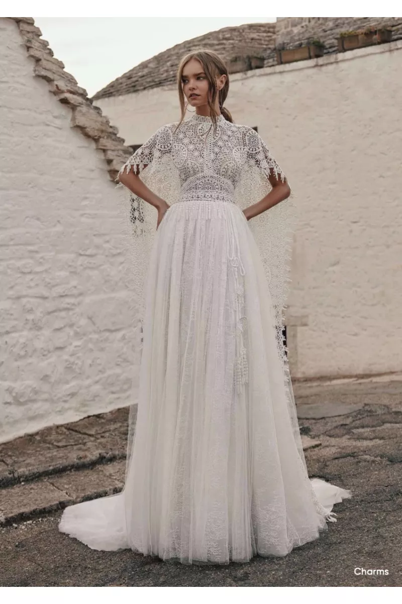 Abito da sposa boho Ariamo Charms in pizzo bianco con dettagli intricati, sfondo architettura rustica.