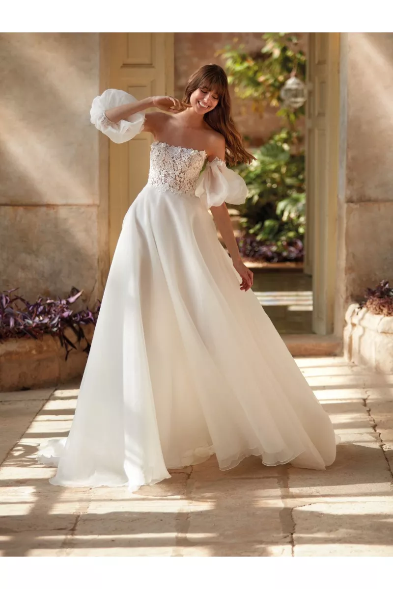 Abito da sposa NASRIN con corpetto in pizzo e maniche voluminose, ambientato in un giardino soleggiato.