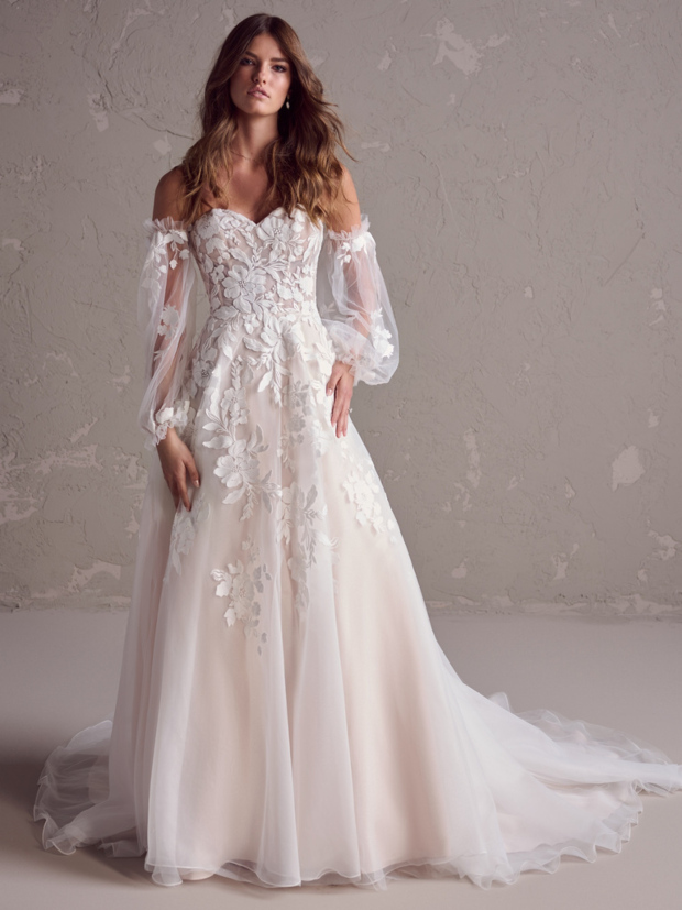 Abito da sposa Ruby Maggie Sottero con maniche trasparenti e dettagli floreali su sfondo neutro.