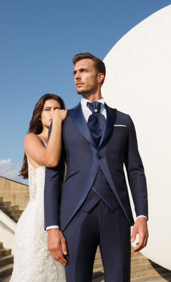 Uomo in abito elegante blu PALAZZO C8 165, donna in abito bianco, sfondo architettonico moderno.