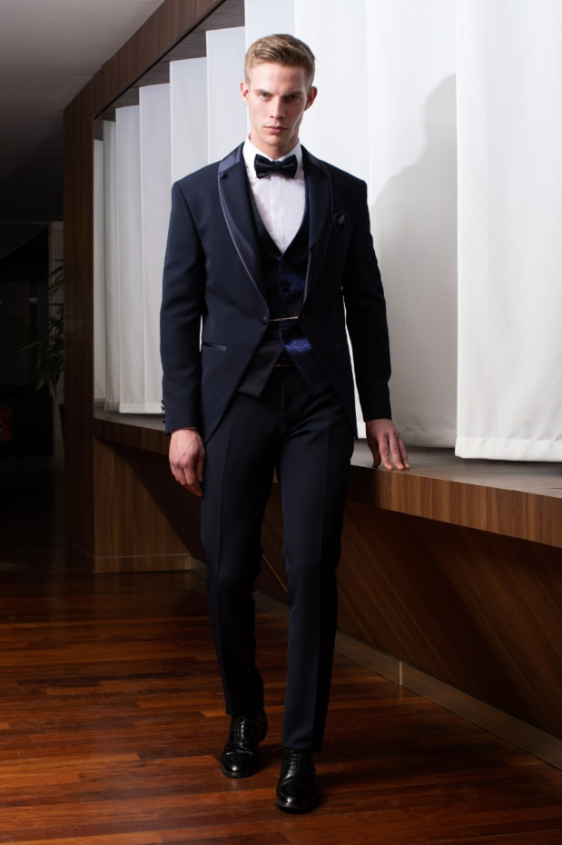 Abito da uomo elegante blu scuro con papillon, camicia bianca e scarpe nere, su sfondo moderno in legno.