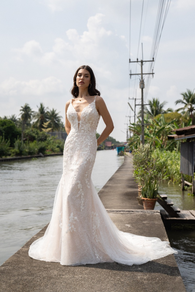 Abito da sposa Amelie Deluxe 10107 con dettagli floreali, sfondo tropicale e cielo nuvoloso.