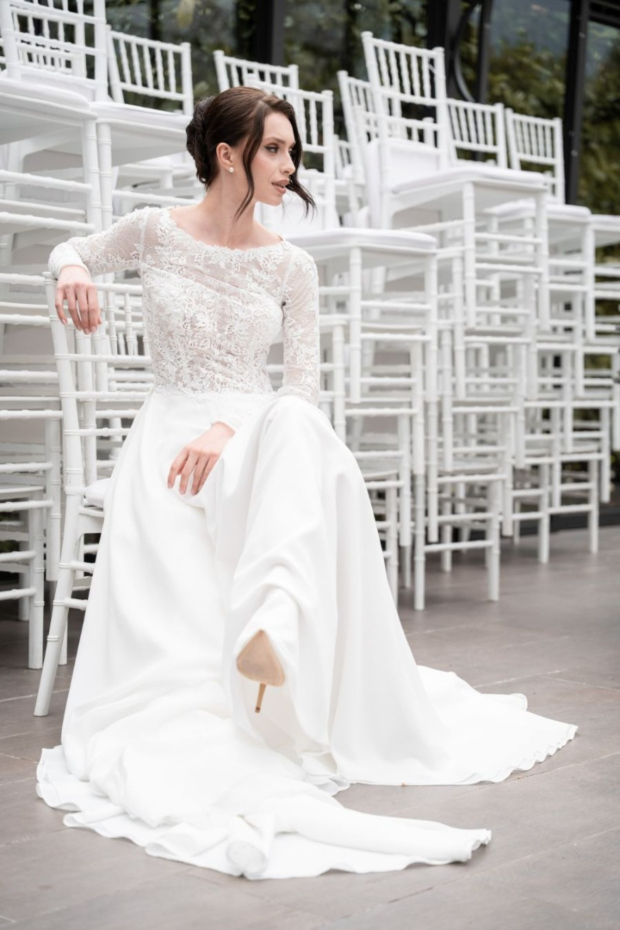 Abito da sposa bianco con pizzo, modello TAISSA, collezione The Romantica, donna seduta tra sedie bianche.