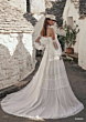 Abito da sposa boho Arabella, maniche trasparenti, sfondo di case in pietra.