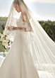 Abito da sposa Lyla St. Patrick con velo in pizzo floreale, sfondo naturale luminoso.