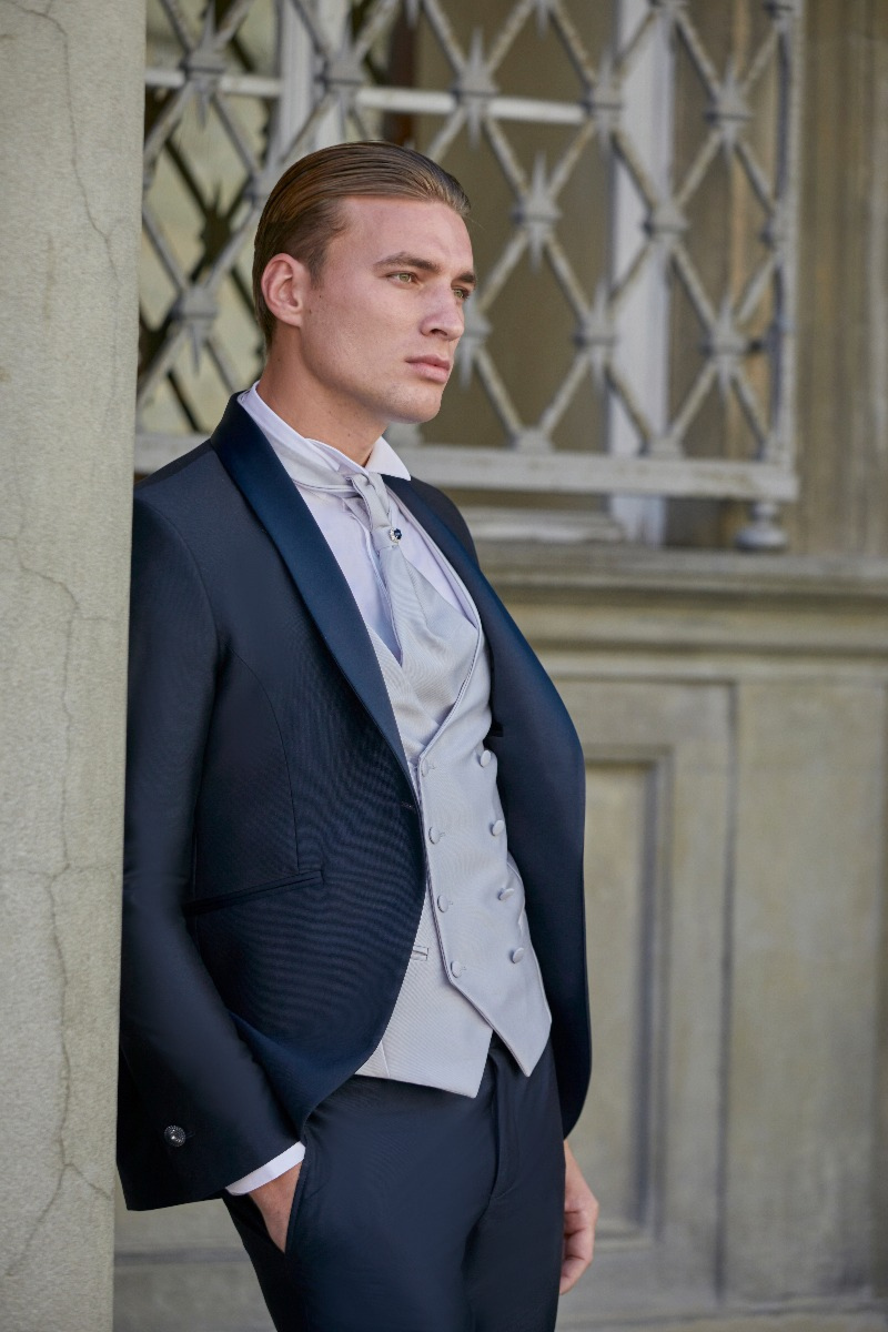 Abito elegante uomo Rocchini 11, giacca scura e gilet chiaro, indossato da modello davanti a finestra decorata.