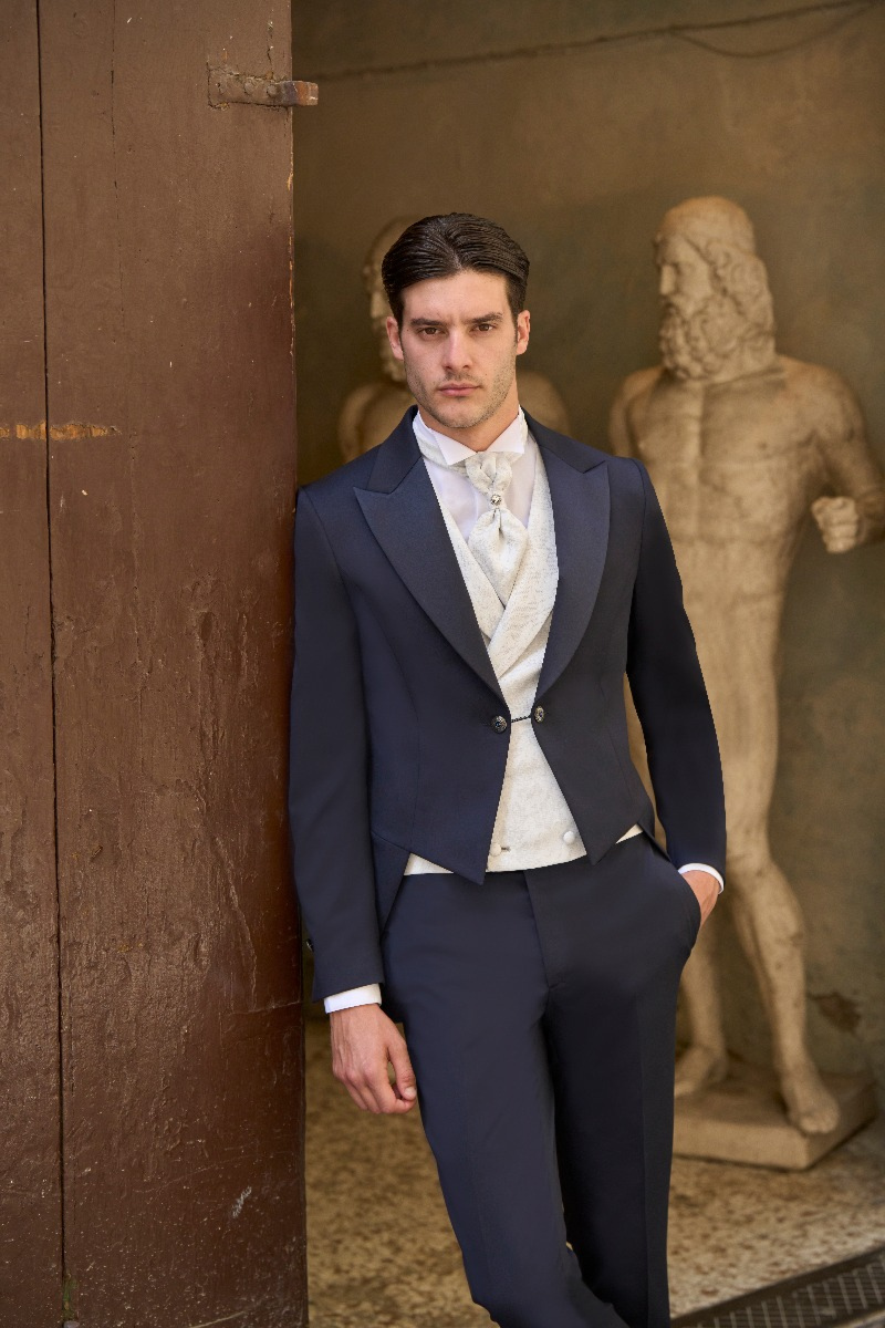 Abito da cerimonia Rocchini 2, uomo in giacca blu e gilet bianco, sfondo con statue antiche.
