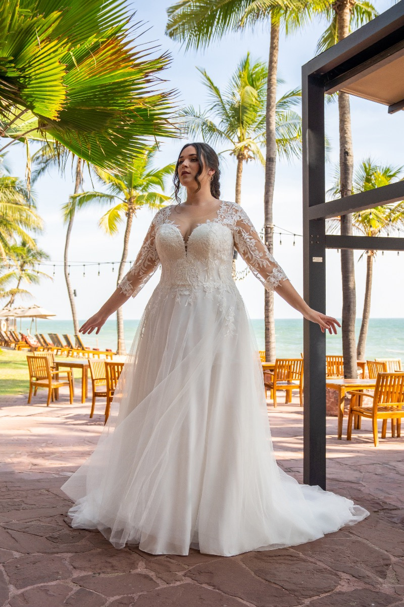 Abito da sposa Bella by Amelie con pizzo e maniche lunghe, ambientato in un giardino tropicale con palme.