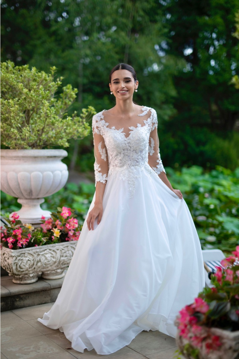 Abito da sposa in pizzo bianco con maniche lunghe, eco linea Sposae Atelier, giardino fiorito sullo sfondo.