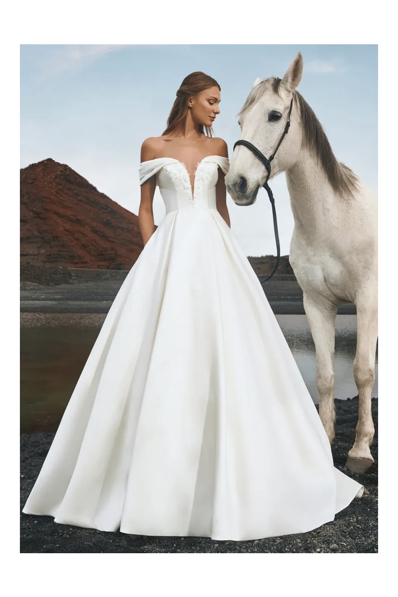Abito da sposa IGUAZU bianco con scollo a cuore, donna accanto a cavallo, paesaggio naturale sullo sfondo.