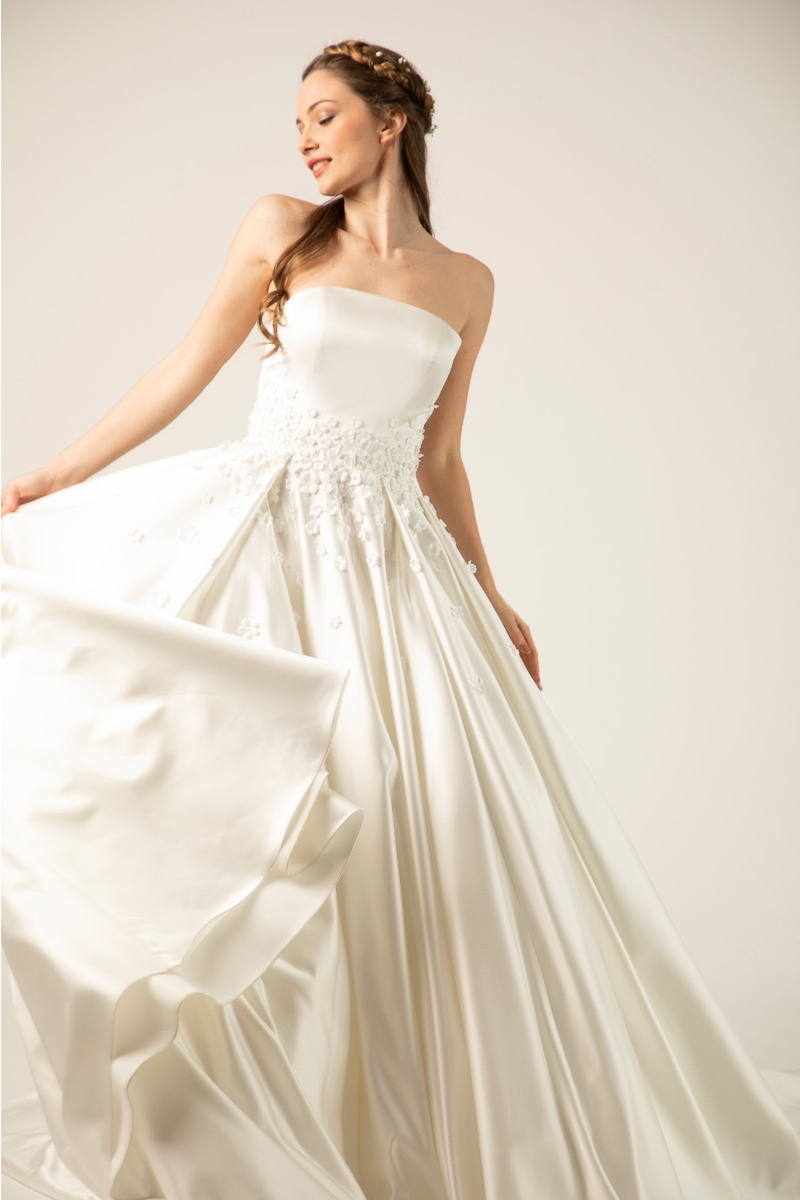 Abito da sposa Monoi in raso bianco, senza spalline, con dettagli floreali, indossato da modella sorridente.