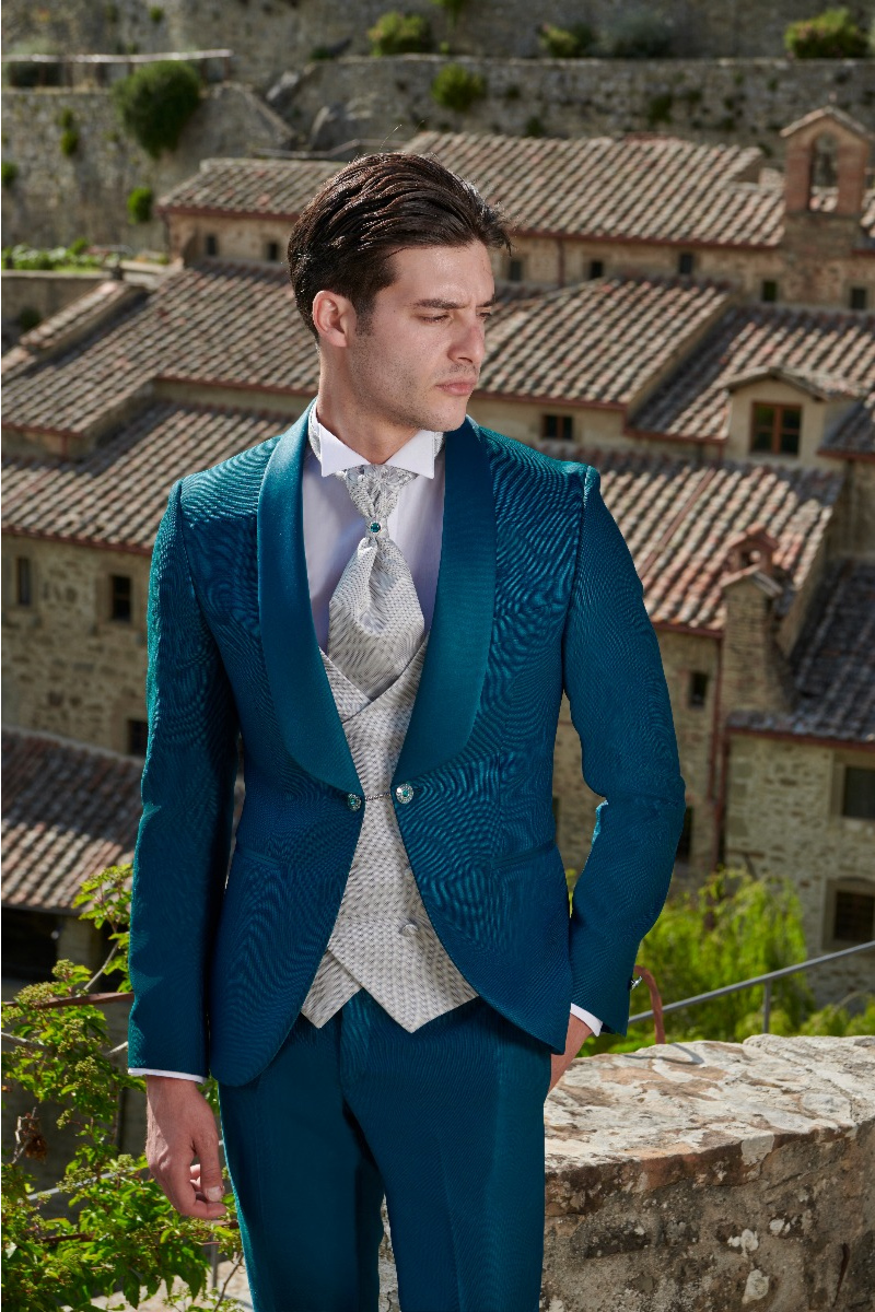Abito elegante Rocchini 9 blu, giacca e cravatta argento, sfondo di tetti in pietra.