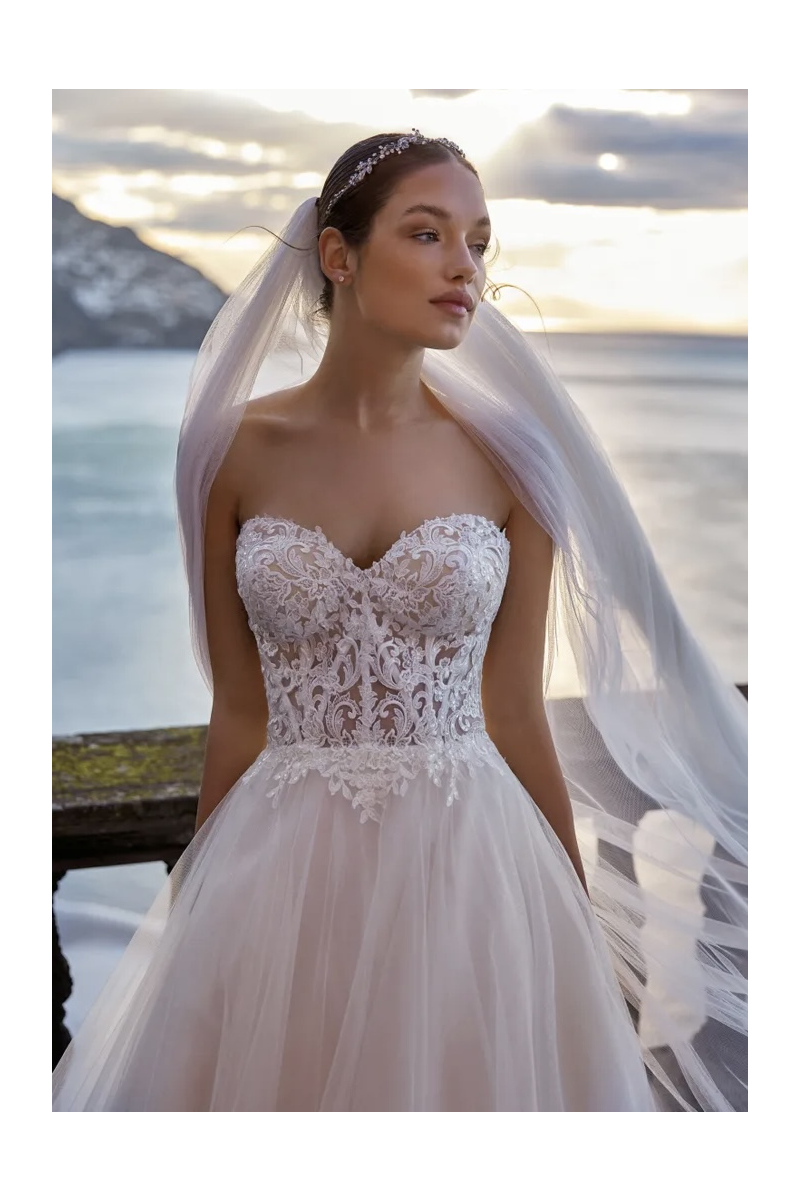 Abito da sposa Nikiya Nicole Jolies, design elegante con corpetto in pizzo e tulle, sfondo paesaggio al tramonto.