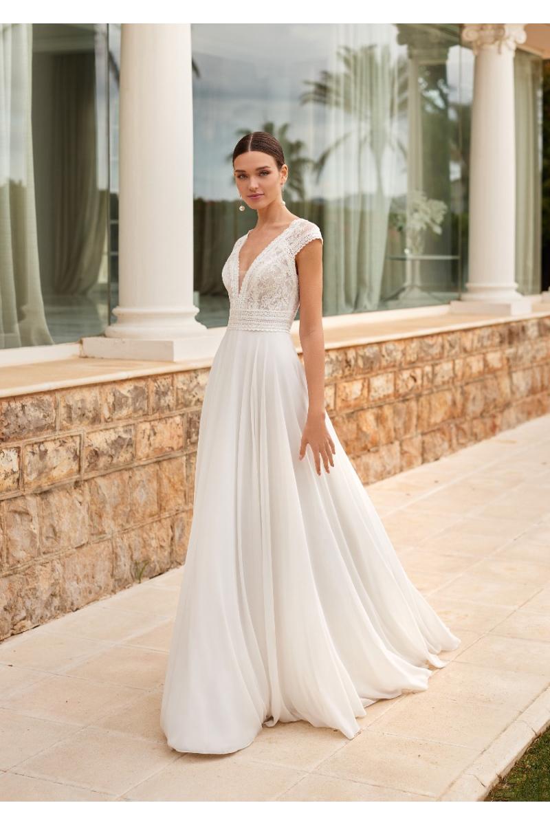 Abito da sposa ALMIA in pizzo bianco con scollo a V, sfondo architettonico elegante.
