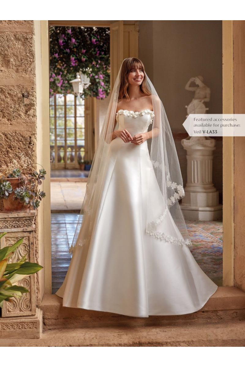 Abito da sposa ALYVIA in raso bianco con velo decorato, ambientato in un elegante scenario rustico.