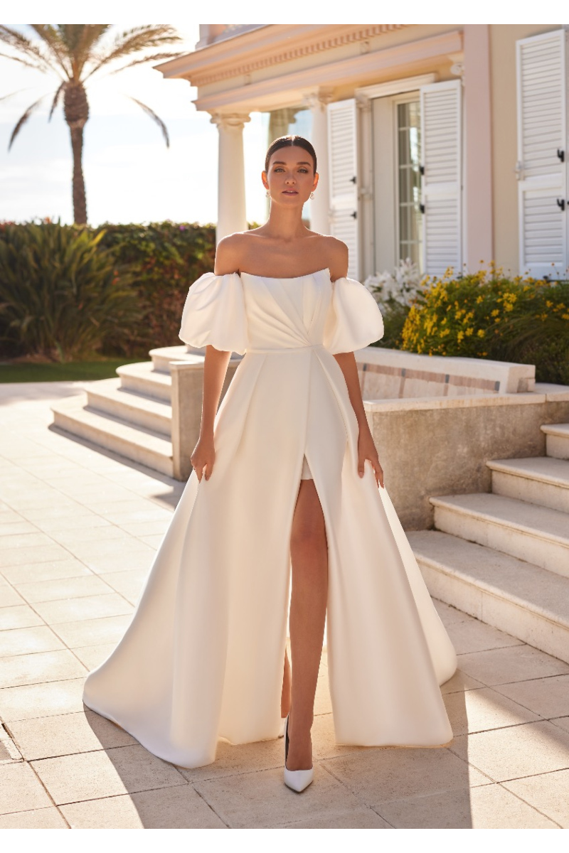 Abito da sposa ANGLESEY con spalle scoperte e spacco, ambientato in un elegante giardino con palme.