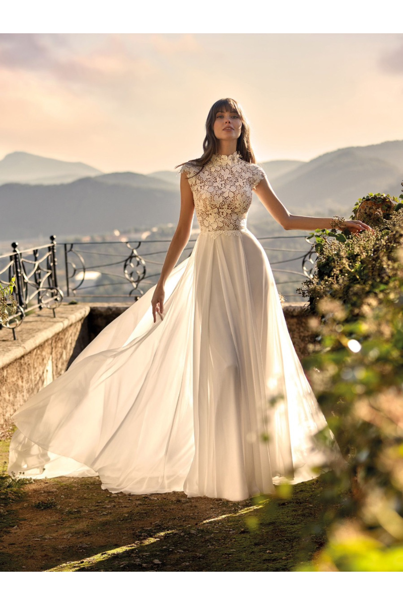 Abito da sposa ANTHA Nicole Colet in pizzo e chiffon, sfondo paesaggio montano al tramonto.