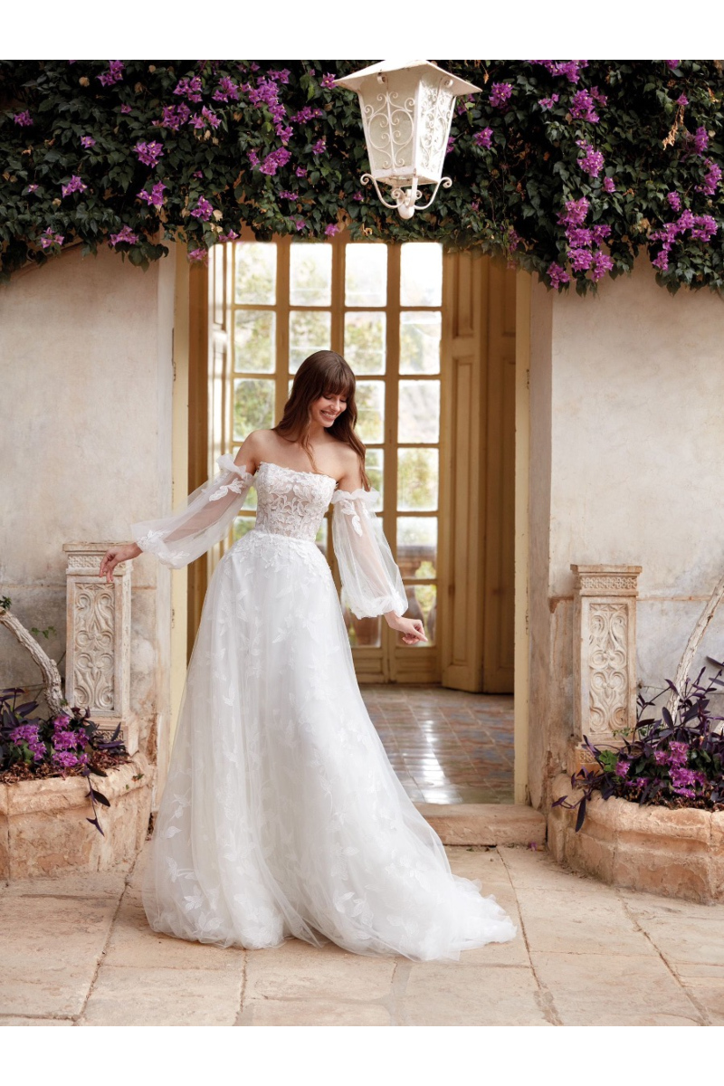 Abito da sposa Aveline con corpetto in pizzo e maniche trasparenti, ambientazione floreale romantica.