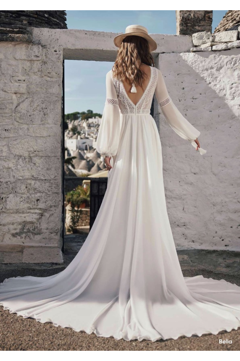 Abito da sposa boho Bella Ariamo, maniche lunghe in pizzo, sfondo architettura bianca.