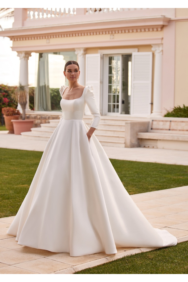 Abito da sposa Bertha in raso bianco con maniche lunghe, scollo quadrato, ambientato in giardino elegante.