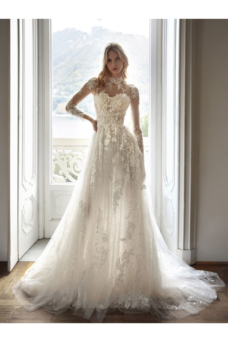 Abito da sposa Nicole Milano CANAIMA in pizzo floreale, maniche lunghe trasparenti, sfondo montano.