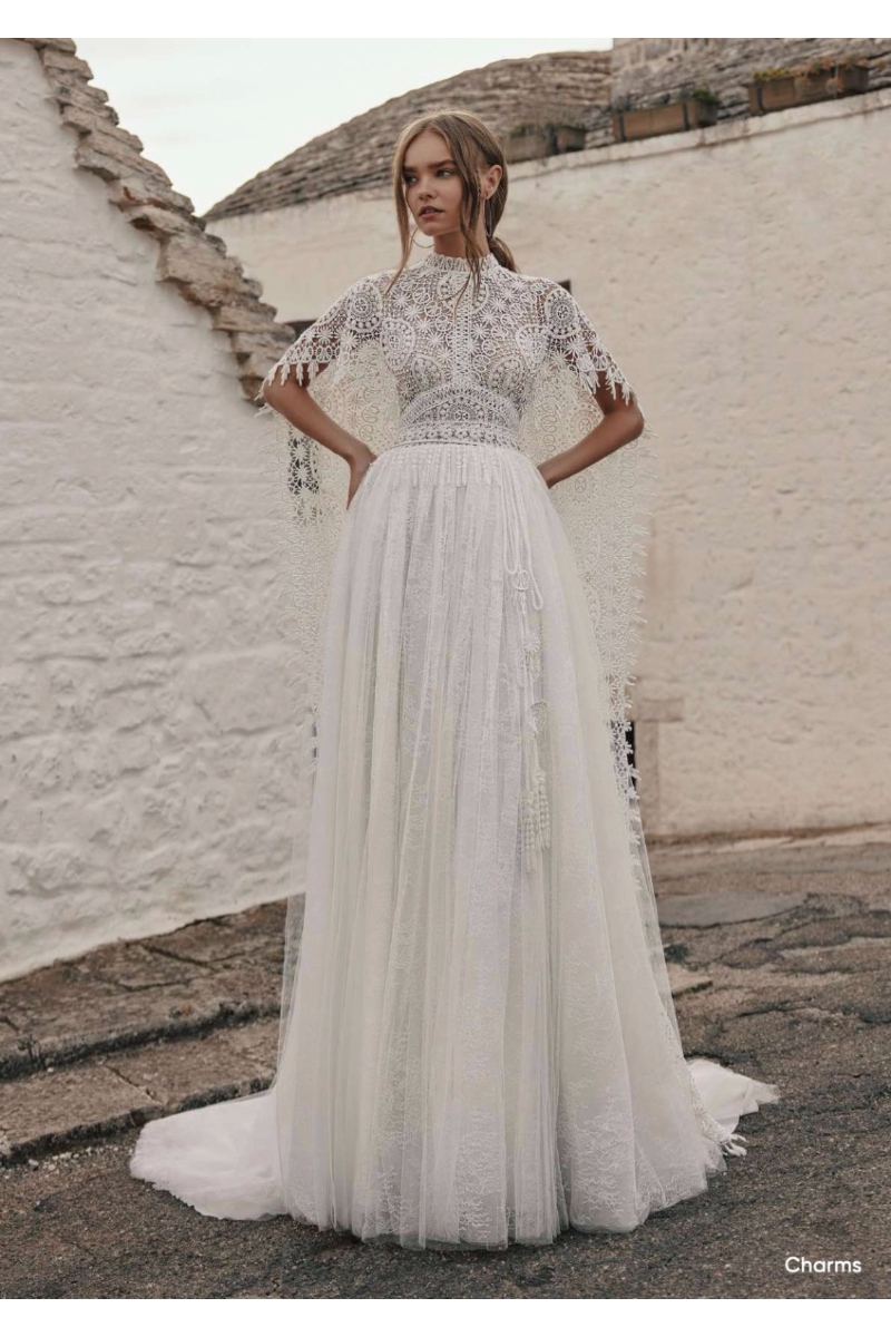 Abito da sposa boho Ariamo Charms in pizzo bianco con dettagli intricati, sfondo architettura rustica.