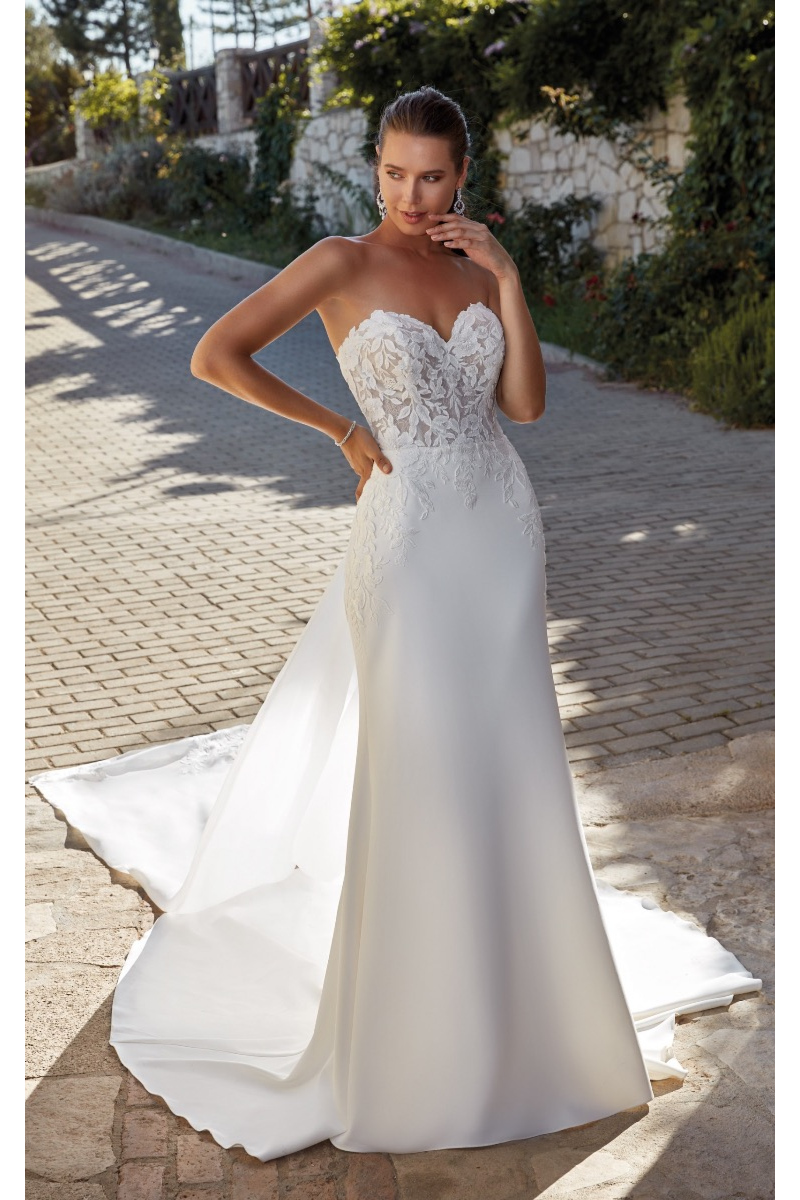 Abito da sposa Cleo di Eddy K con corpetto in pizzo e gonna lunga, ambientato all'aperto su una strada acciottolata.
