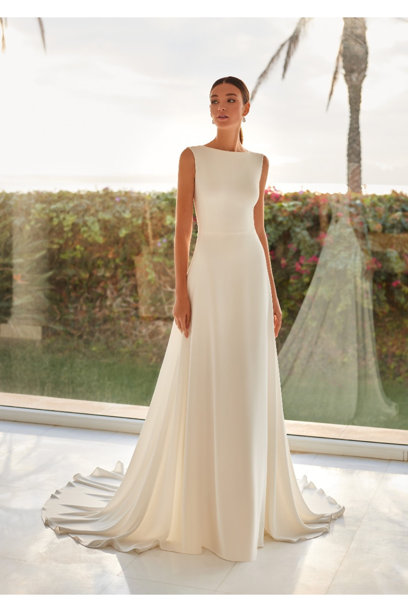 Abito da sposa elegante CORA con design minimalista e scollo a barca, sfondo naturale con palme.