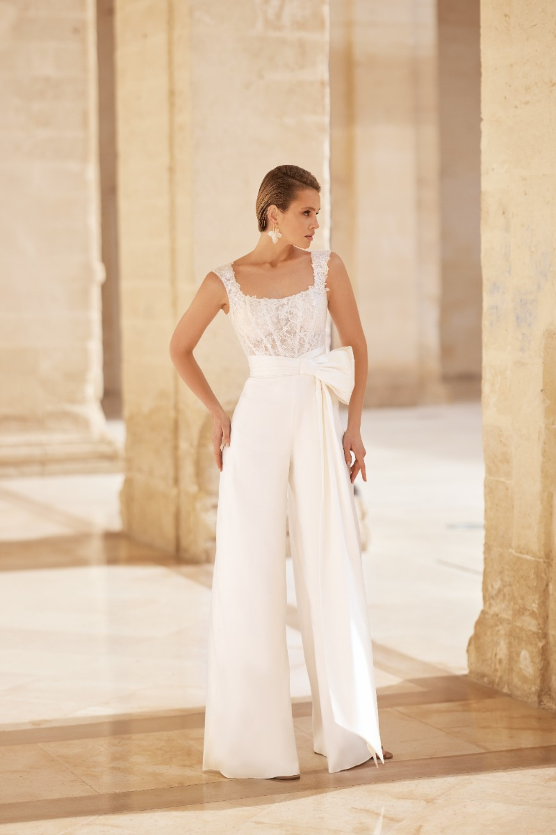 Tuta bianca elegante con dettagli in pizzo, design CORAL di Mark Loren, ambientata in un interno luminoso.