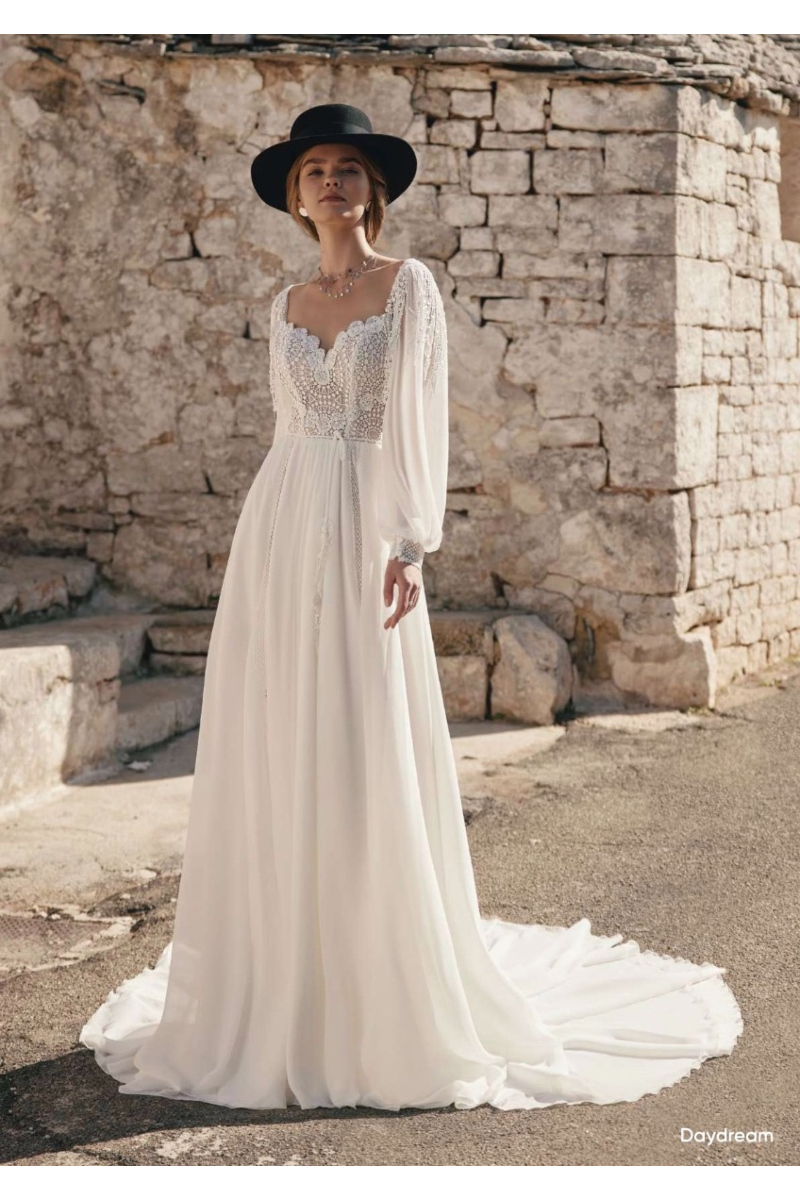 Abito da sposa boho DAYDREAM ARIAMO con maniche lunghe e cappello nero, sfondo rustico in pietra.