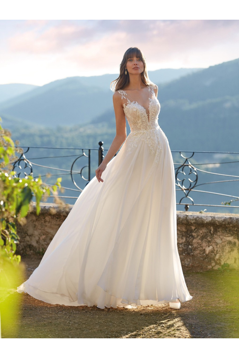 Abito da sposa DEFNE di Nicole Colet, scollo a cuore e dettagli floreali, sfondo montano.
