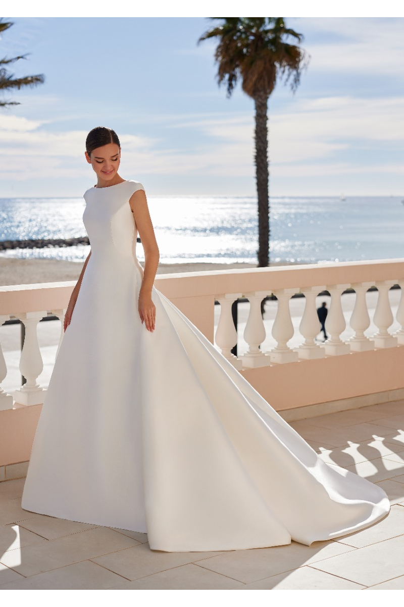 Abito da sposa Doriana, elegante e classico, in riva al mare con sfondo di palme e cielo azzurro.