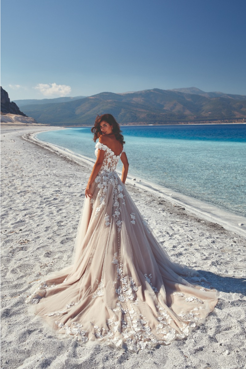Abito da sposa Lima Eddy K in spiaggia, dettagli floreali, mare e monti sullo sfondo.