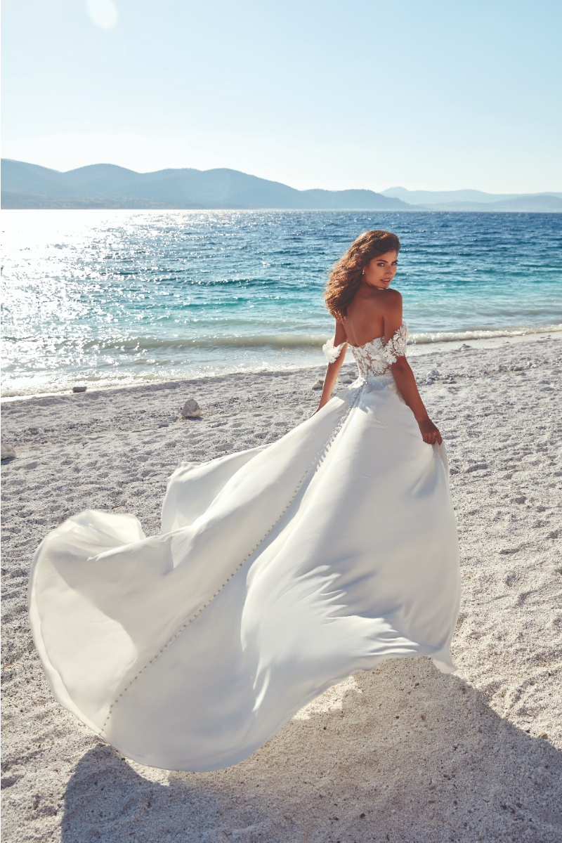 Abito da sposa YARA di Eddy K su spiaggia, donna di spalle, mare azzurro sullo sfondo.