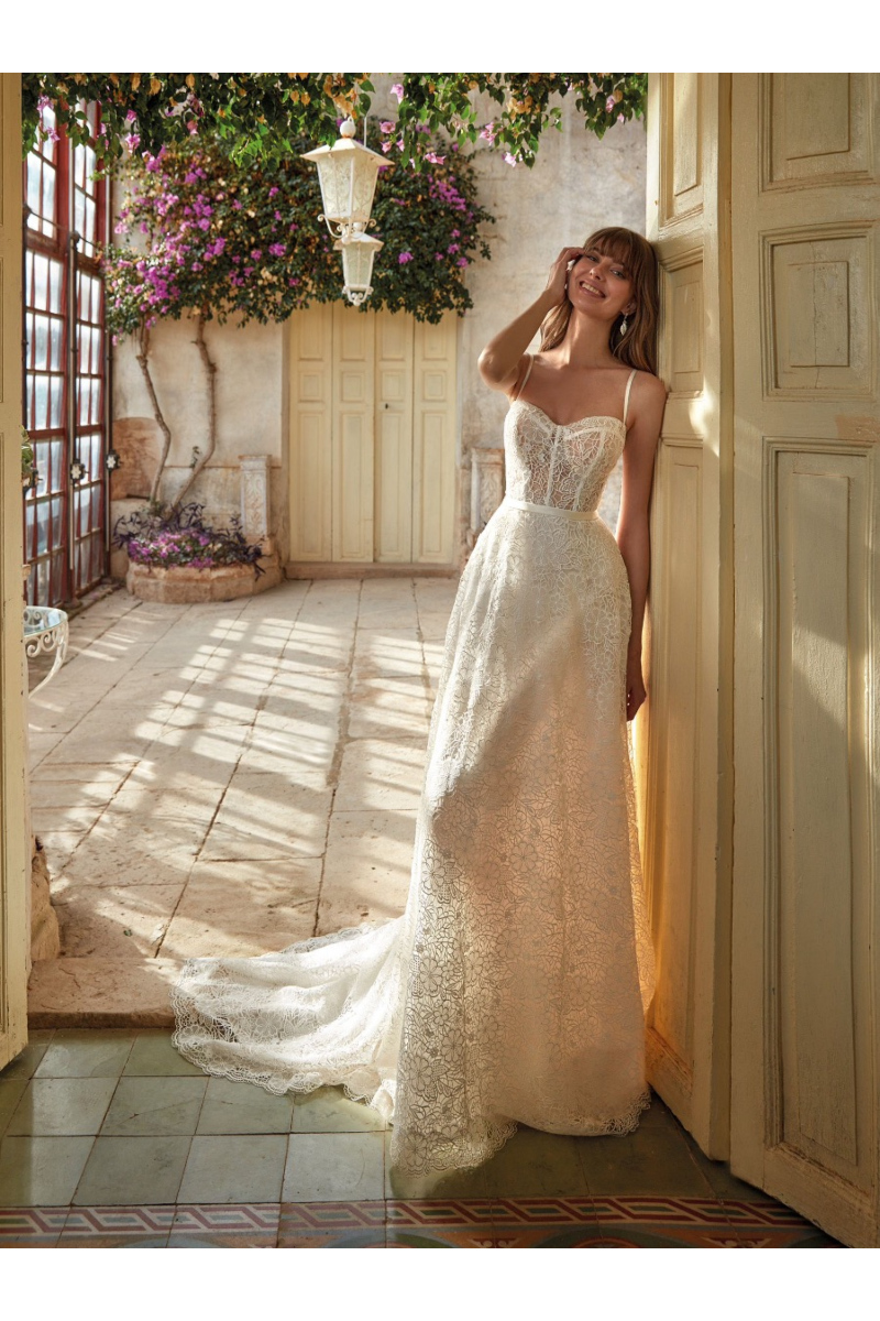 Abito da sposa EILA in pizzo bianco, ambientato in un cortile fiorito con luce naturale.
