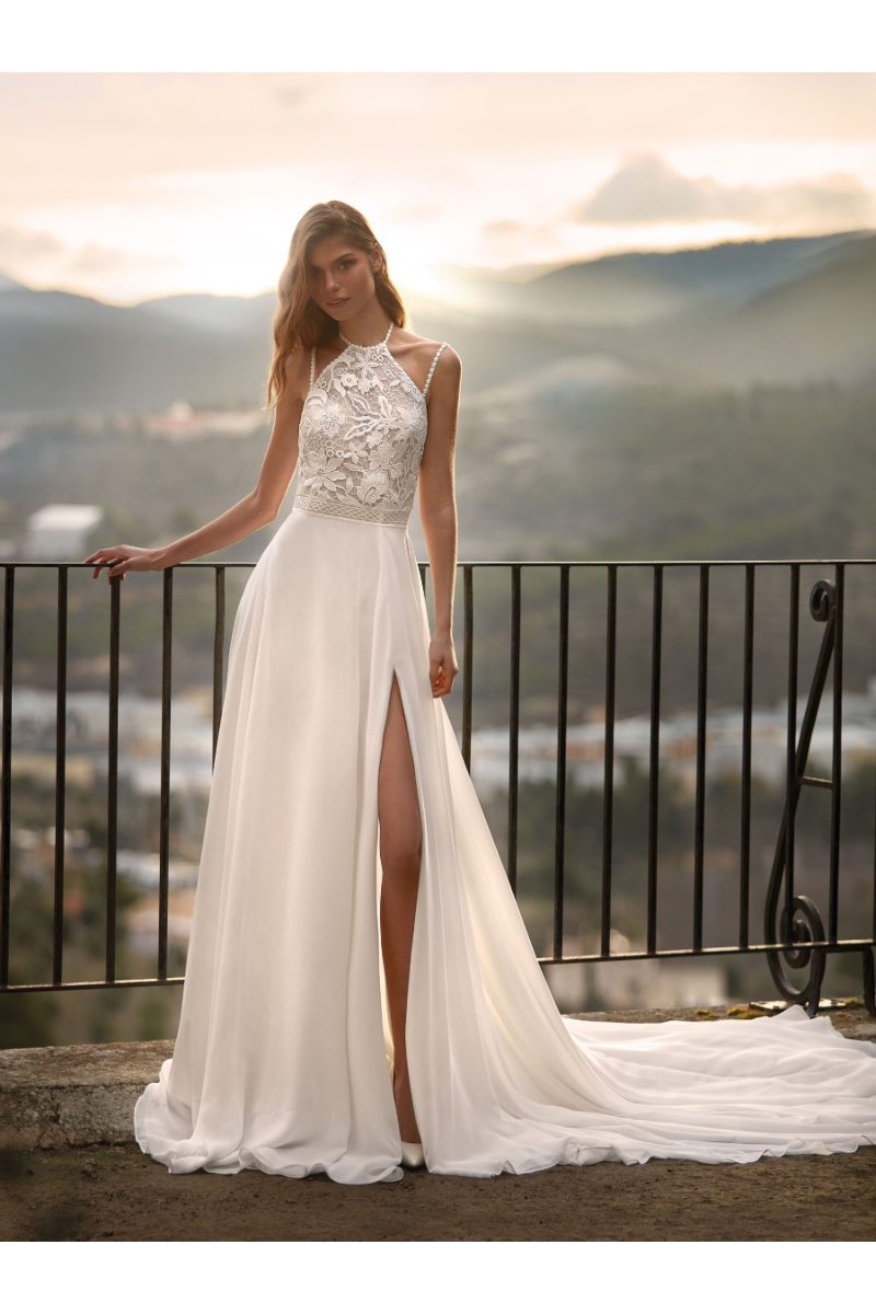 Abito da sposa ELOWYN Nicole Colet con corpetto in pizzo e gonna con spacco, sfondo panoramico al tramonto.