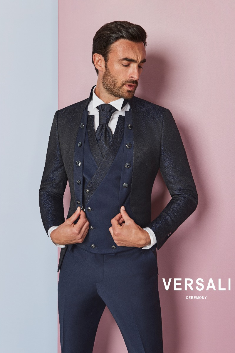 Abito da cerimonia uomo FIT 31 Versali, giacca blu elegante con bottoni, su sfondo rosa e azzurro.