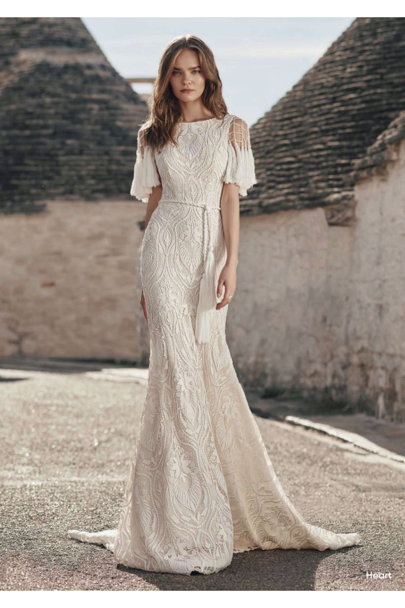 Abito da sposa boho ricamato, maniche a rete, sfondo architettura tradizionale.